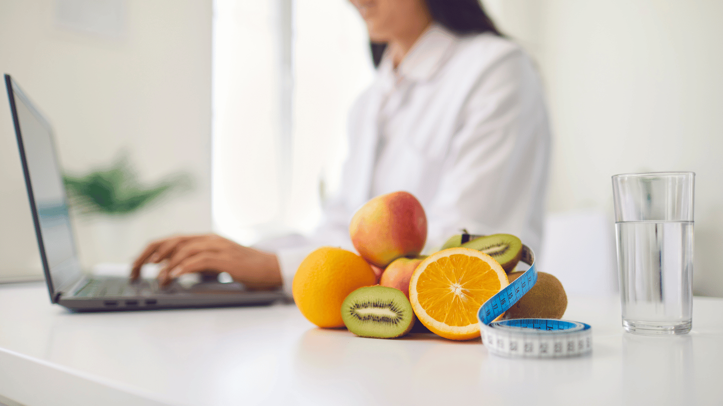 A doctor working on a laptop with fresh fruits, a glass of water, and a measuring tape on the table, symbolizing convenient online weight loss with Mounjaro through telemedicine